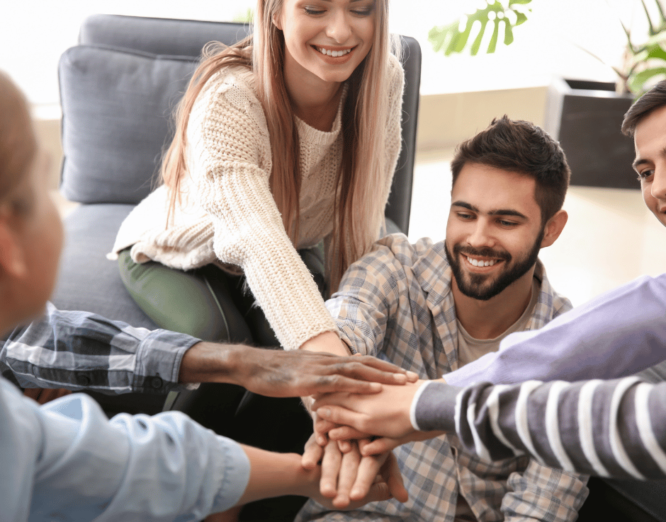 BST-C Fünf Personen sitzen gemeinsam in einem Wohnzimmer und legen ihre Hände übereinander als Zeichen von Teamgeist und Zusammenhalt.