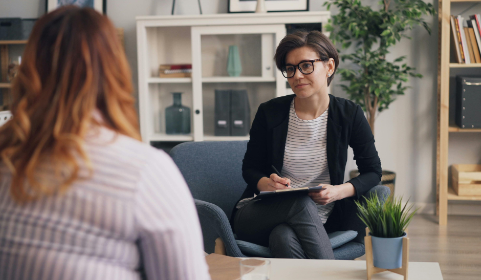Therapiegespräch zwischen zwei Personen in einem modernen Raum, eine Person mit Klemmbrett hört aufmerksam zu.